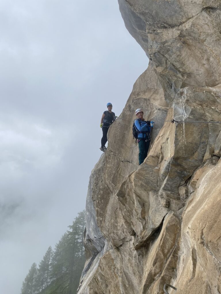 Two climbers in helmets and harnesses standing on a granite cliff face with a fixed cable, cloudy sky and treetops below
