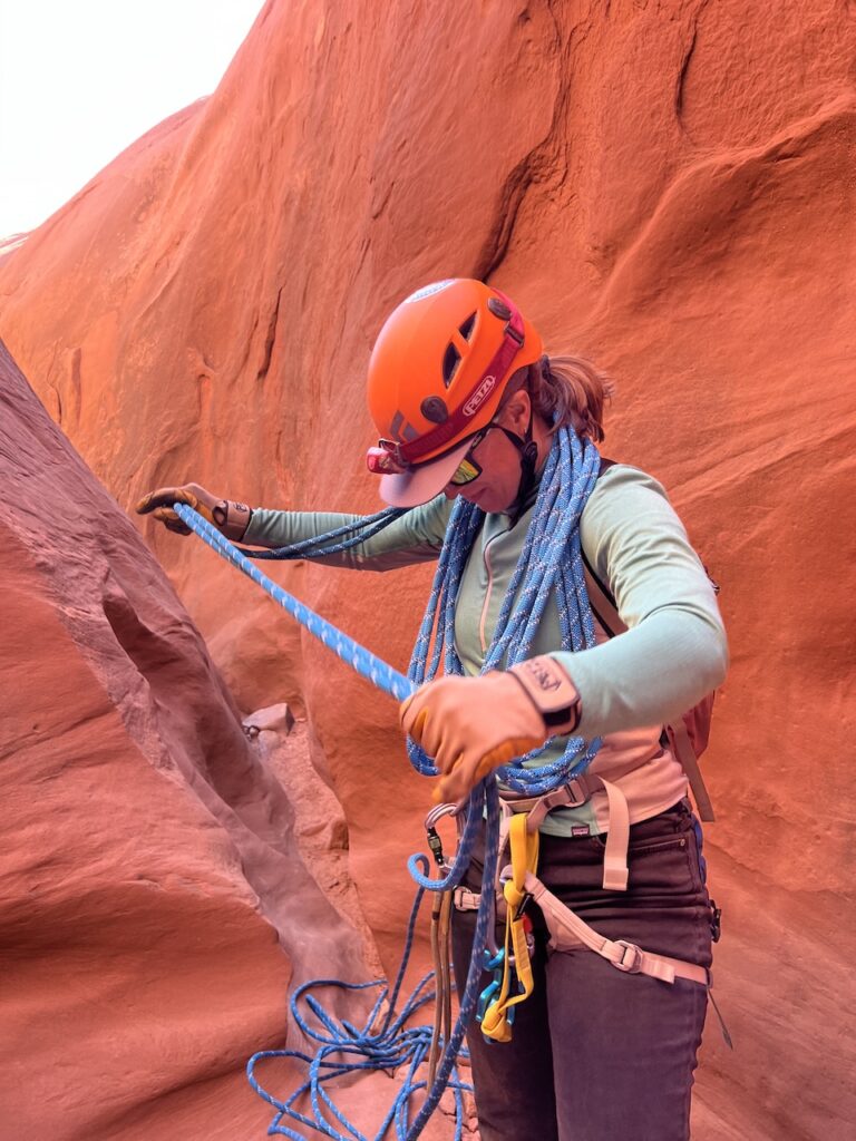 Kaycie Robinson in an orange helmet and harness coiling a blue climbing rope inside a red sandstone canyon slot